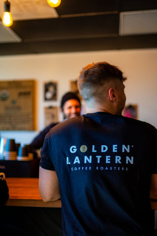 Customer wearing a 'Golden Lantern Coffee Roasters' shirt waiting for a lattee.