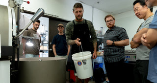 Person holding a bucket of un-roasted beans about to pour into a coffee roasting machine, surrounded by onlookers.
