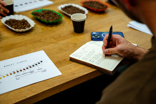 Person taking notes at a coffee tasting event with roasted coffee beans and cups on a table.